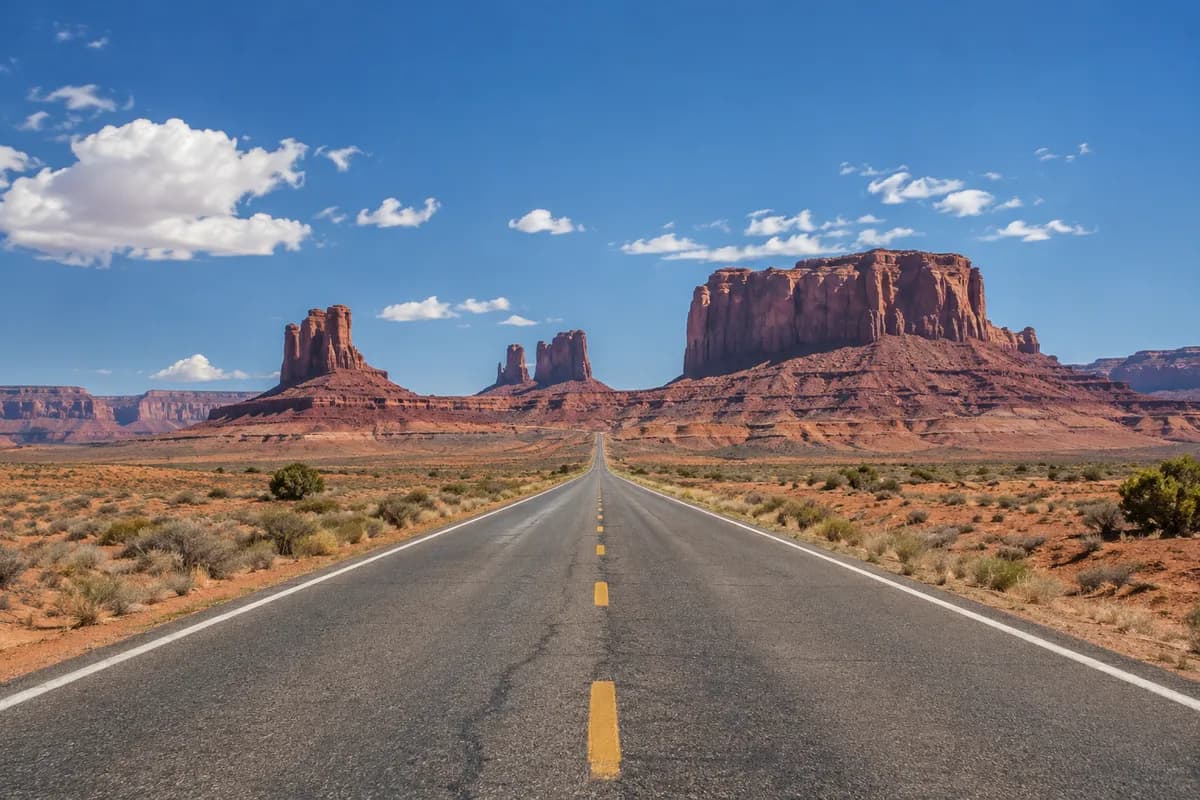 A straight desert highway running toward red sandstone buttes under a midday sky.